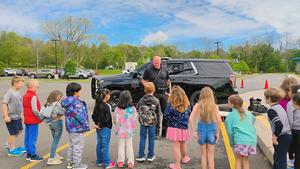 A law enforcement officer speaks with a group of elementary students in front of a patrol vehicle.