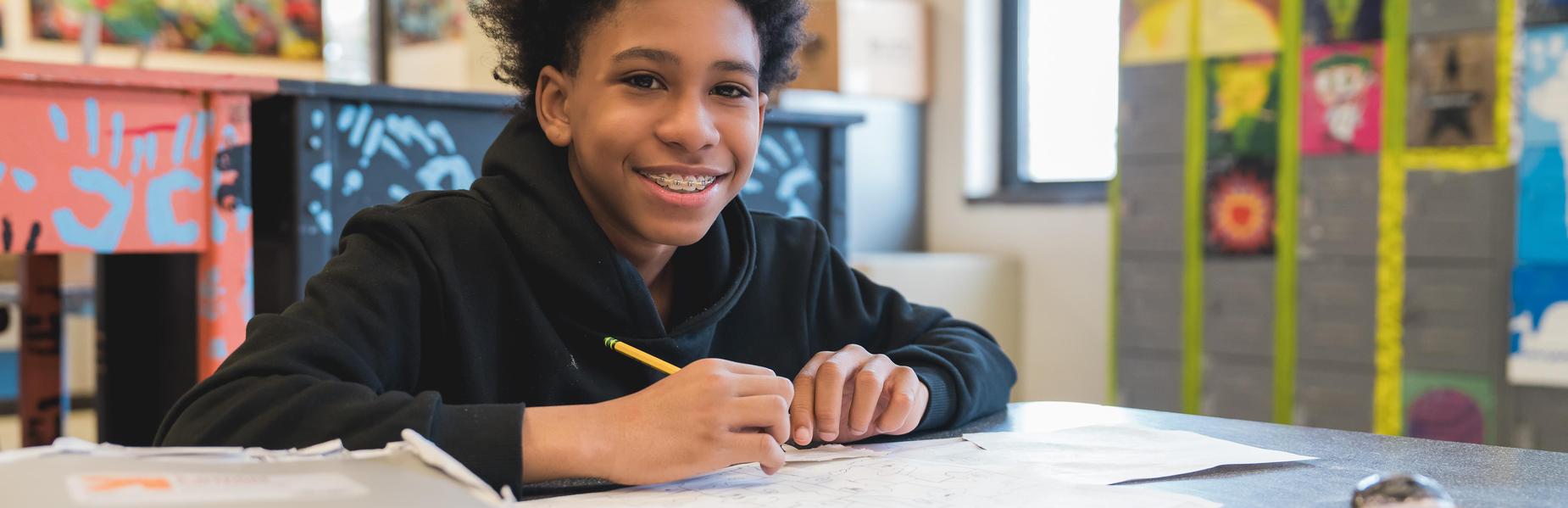 Smiling student working on a drawing at a table filled with art supplies.