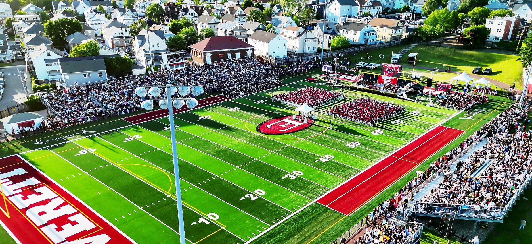 Aerial view of a graduation ceremony on a sports field with crowds gathered.