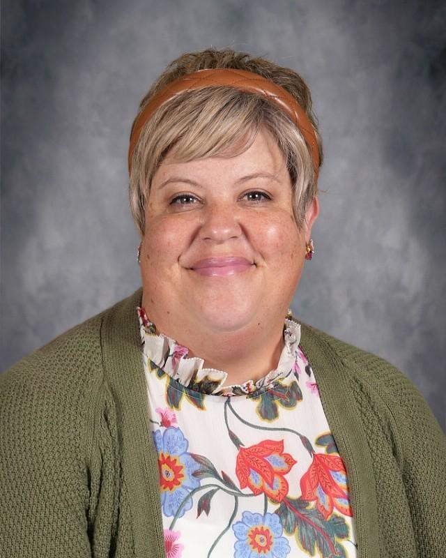 Smiling woman with short hair wearing a patterned blouse against a grey background.