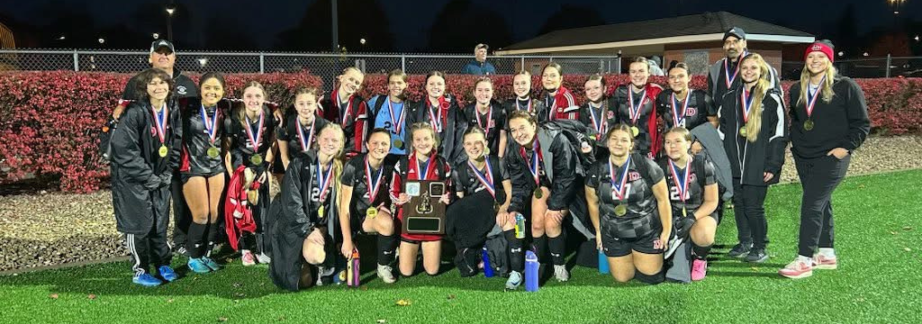 Group photo of the winning girls' soccer team with medals and a trophy on a field at night.