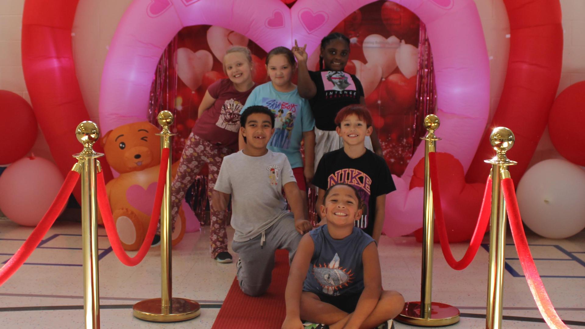 Group of children posing happily in front of a heart-themed decoration.