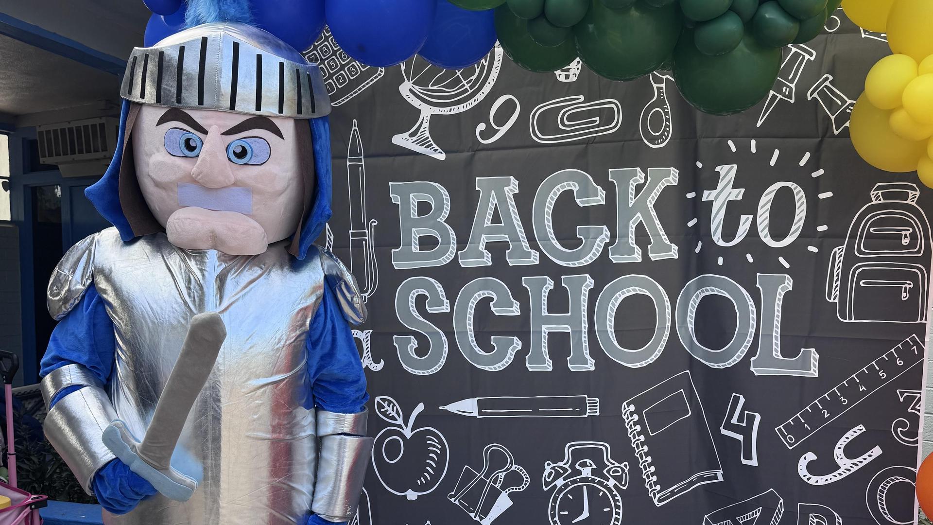 A mascot in a silver costume stands beside a colorful 'Back to School' backdrop.