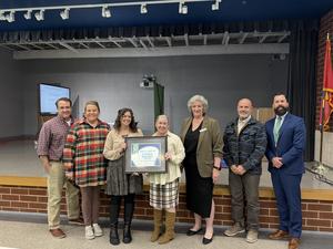 7 adults standing smiling, 2 adults in middle holding plaque of an award