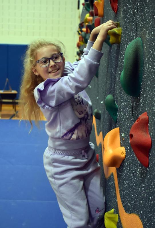 A girl climbs a colorful rock wall in a gym, smiling and wearing glasses.