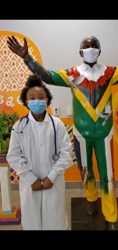 Image shows a scholar dressed in a white lab coat, standing in front of a statue of Nelson Mandela