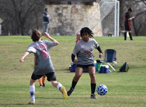 Two students run on a green field with a blue and white soccer ball
