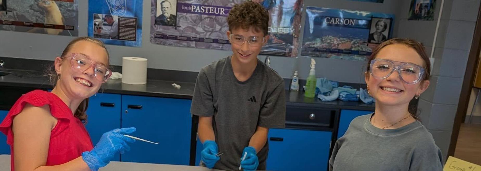 Three students smiling in lab coats working on a science project at a table.
