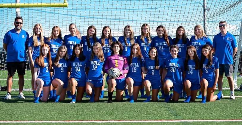 A soccer team of girls in blue uniforms posing together with a coach on the field.