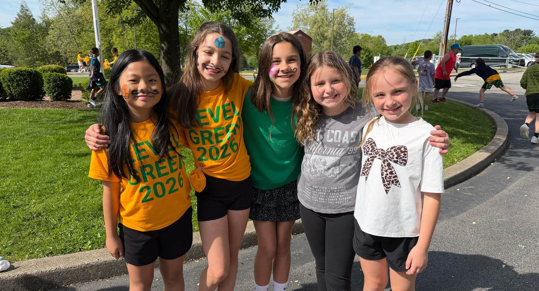 Five young girls stand arm in arm in a parking lot wearing t-shirts and three have moustaches painted on their faces