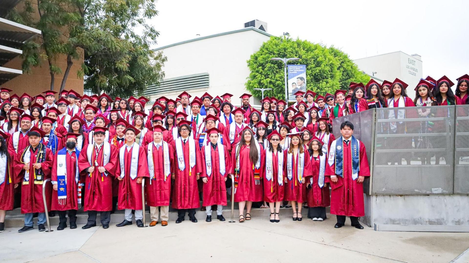 A large group of graduates in red gowns stands together outdoors.