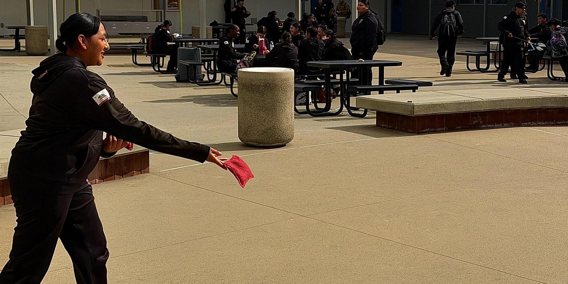 A woman throws a red item while students socialize at tables in a school courtyard.