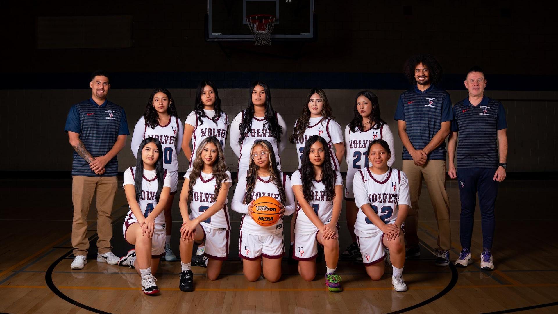 Basketball team in uniforms posed in a gym with a hoop behind them.