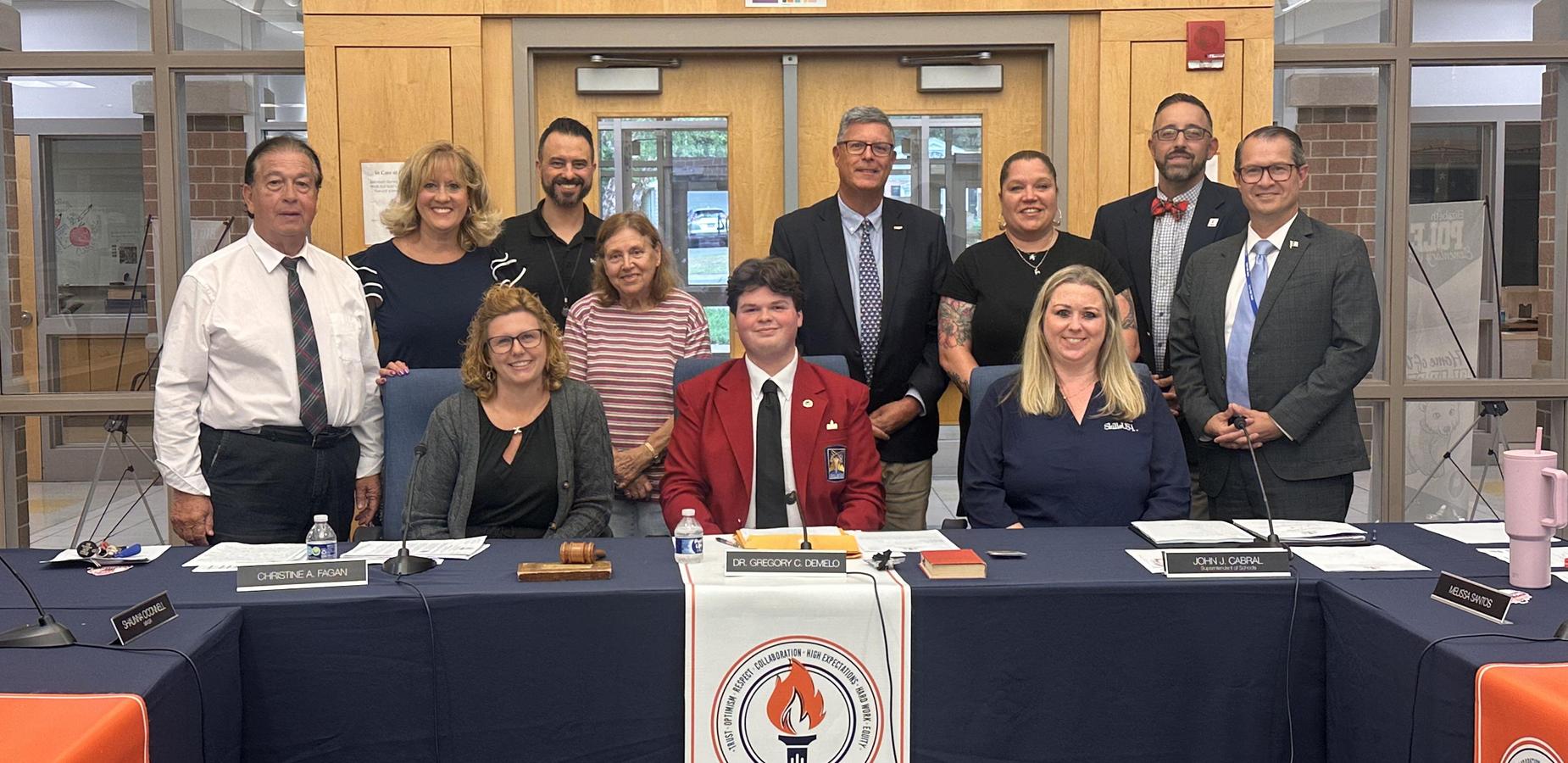 A group of ten adults and one young man in a red blazer stand behind a long table with a blue cloth and a banner that says "TORCHHE." The group is smiling and posing for a photo in what appears to be a public meeting room.
