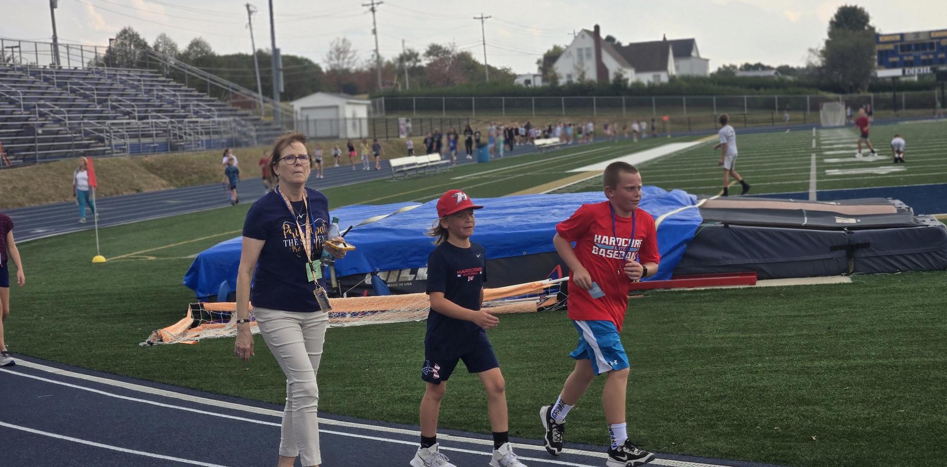 A teacher walking with two boys on a track field near a sports event.