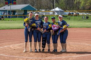 5 softball players dressed in uniforms on field holding flowers