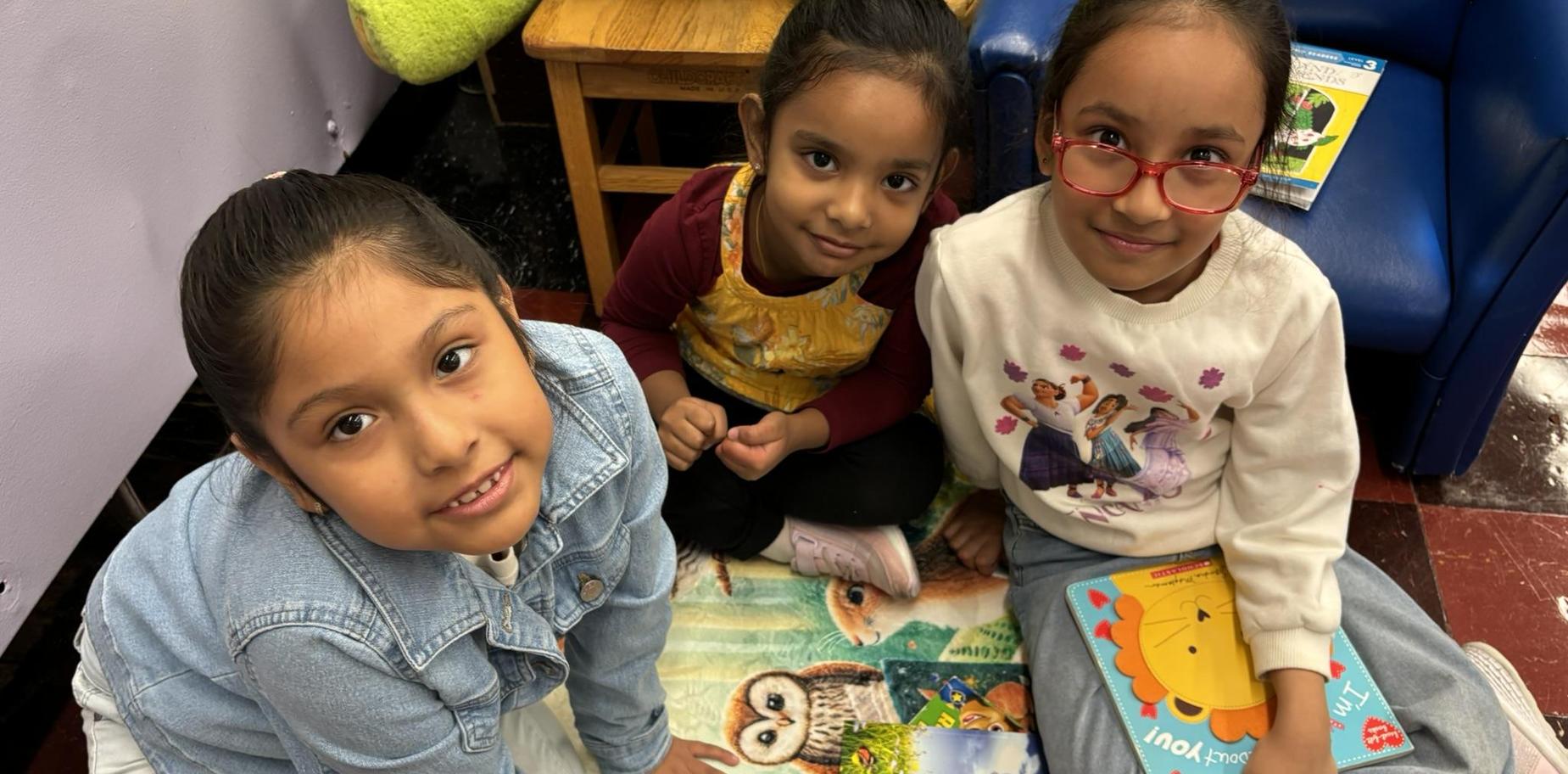Three children sitting on a colorful rug with books around them, smiling at the camera.