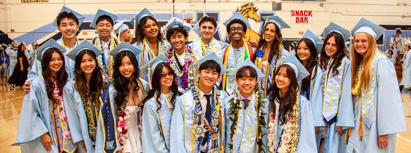 Large group of graduates in blue gowns and leis, smiling together indoors.