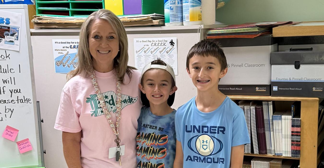 A teacher posing with two boys in casual shirts and shorts, smiling in a classroom.