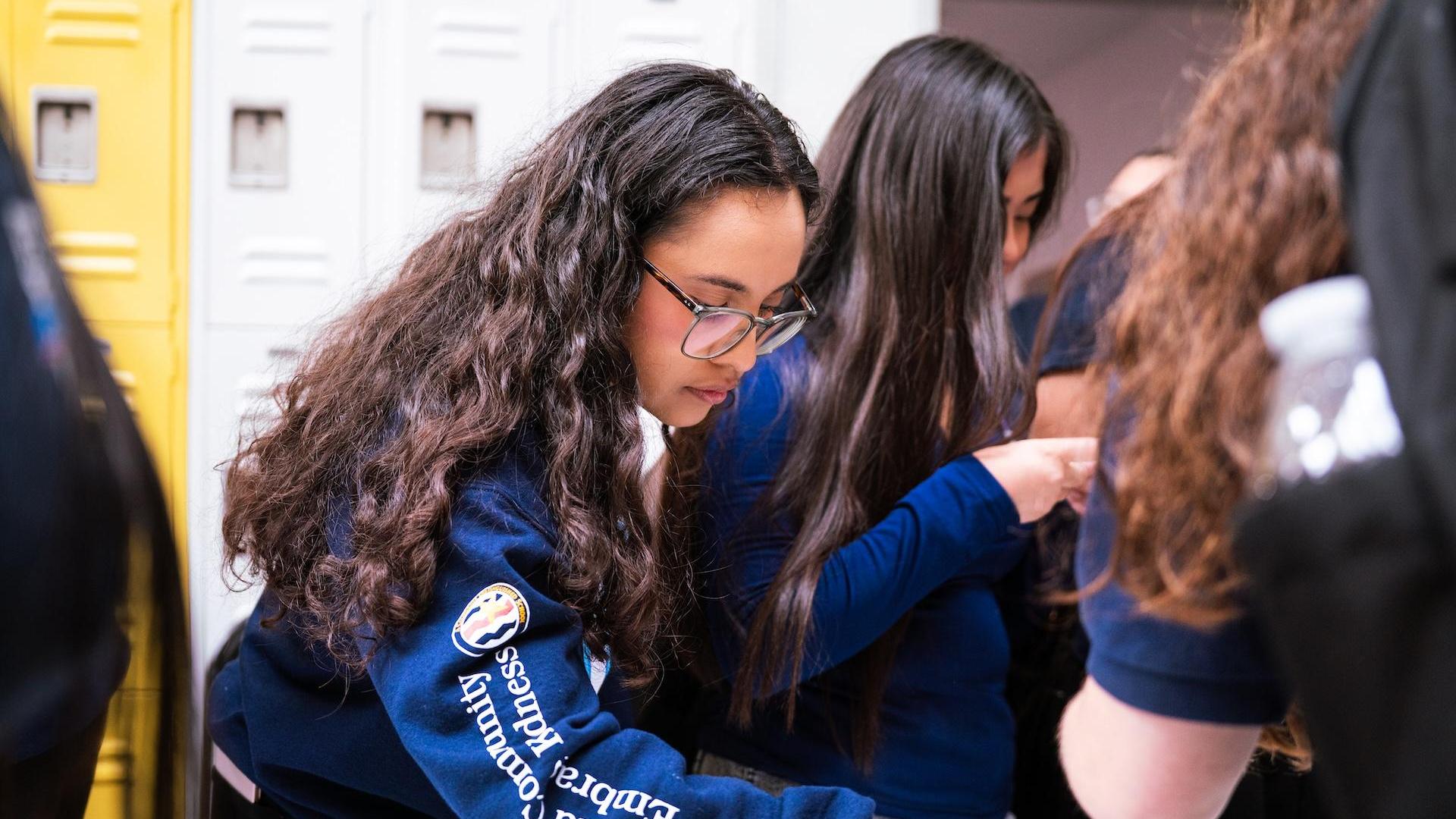 Students interacting near lockers, one girl focused on her work.