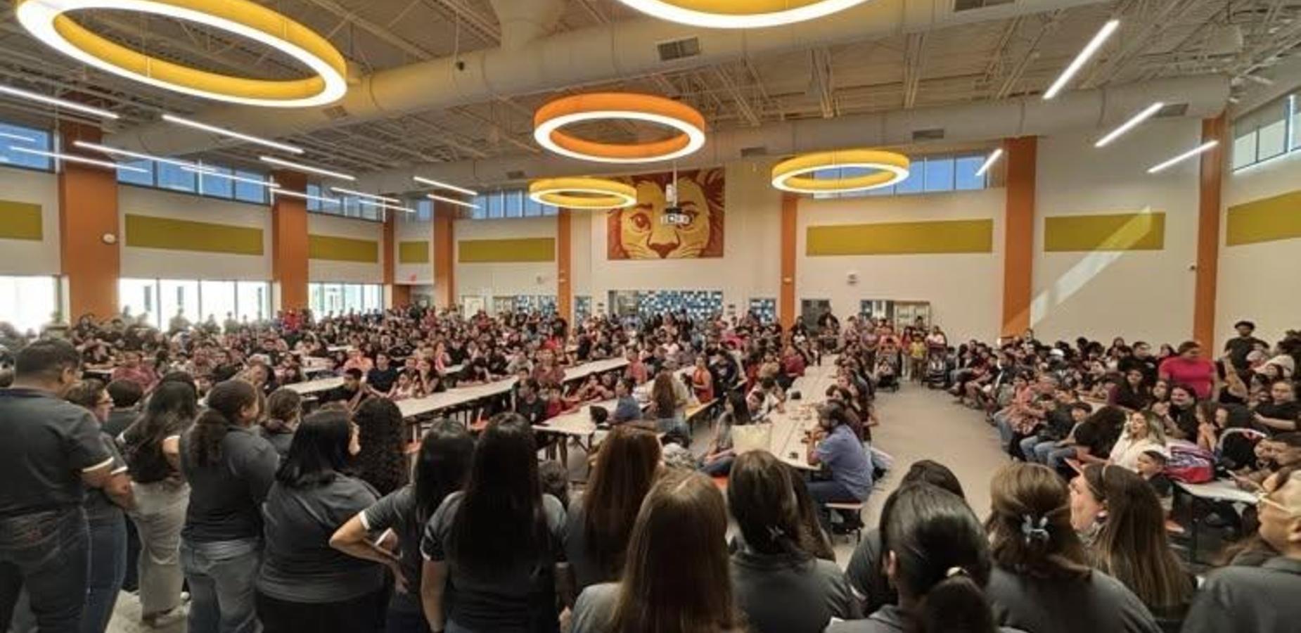 A large crowd in a cafeteria, many seated and participating in an event, with staff members in gray shirts.