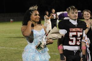 A girl in a blue dress holds flowers next to a football player with a crown.