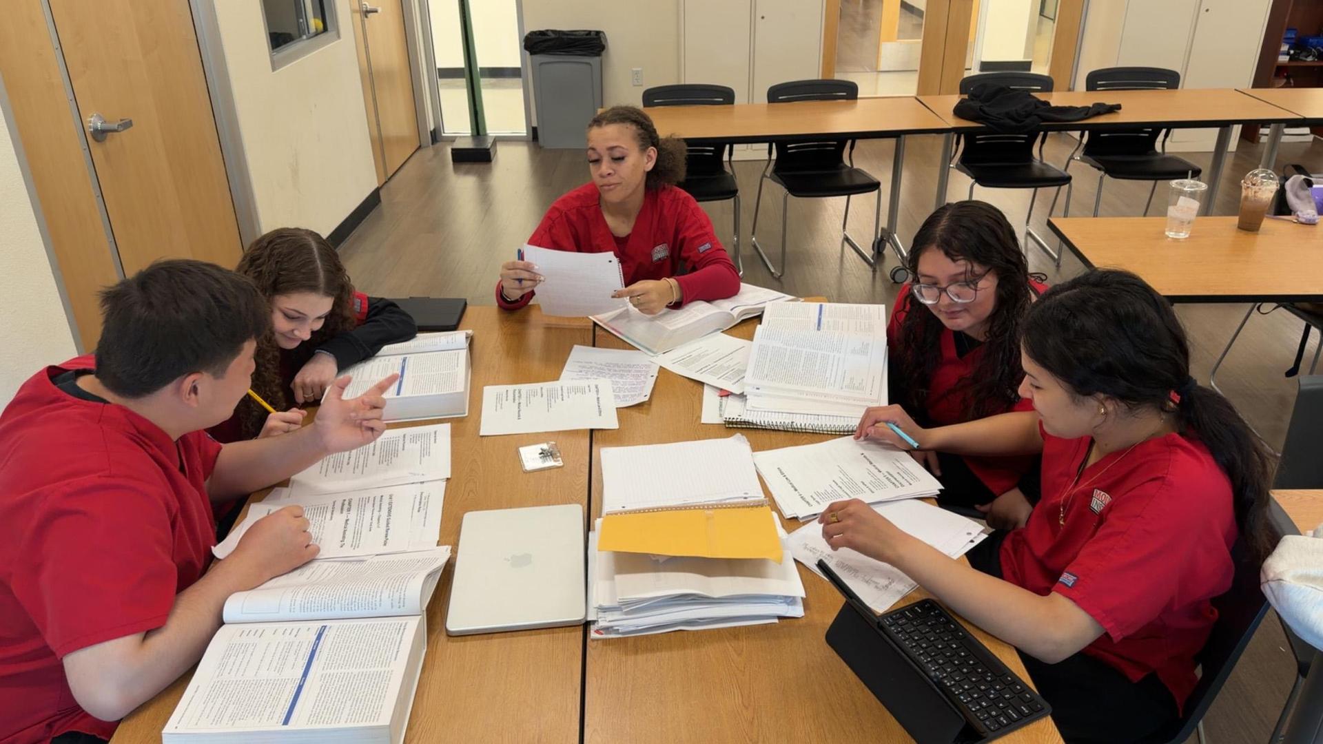 group of medical assisting students in red scrub tops sitting at a table looking at notes and textbooks