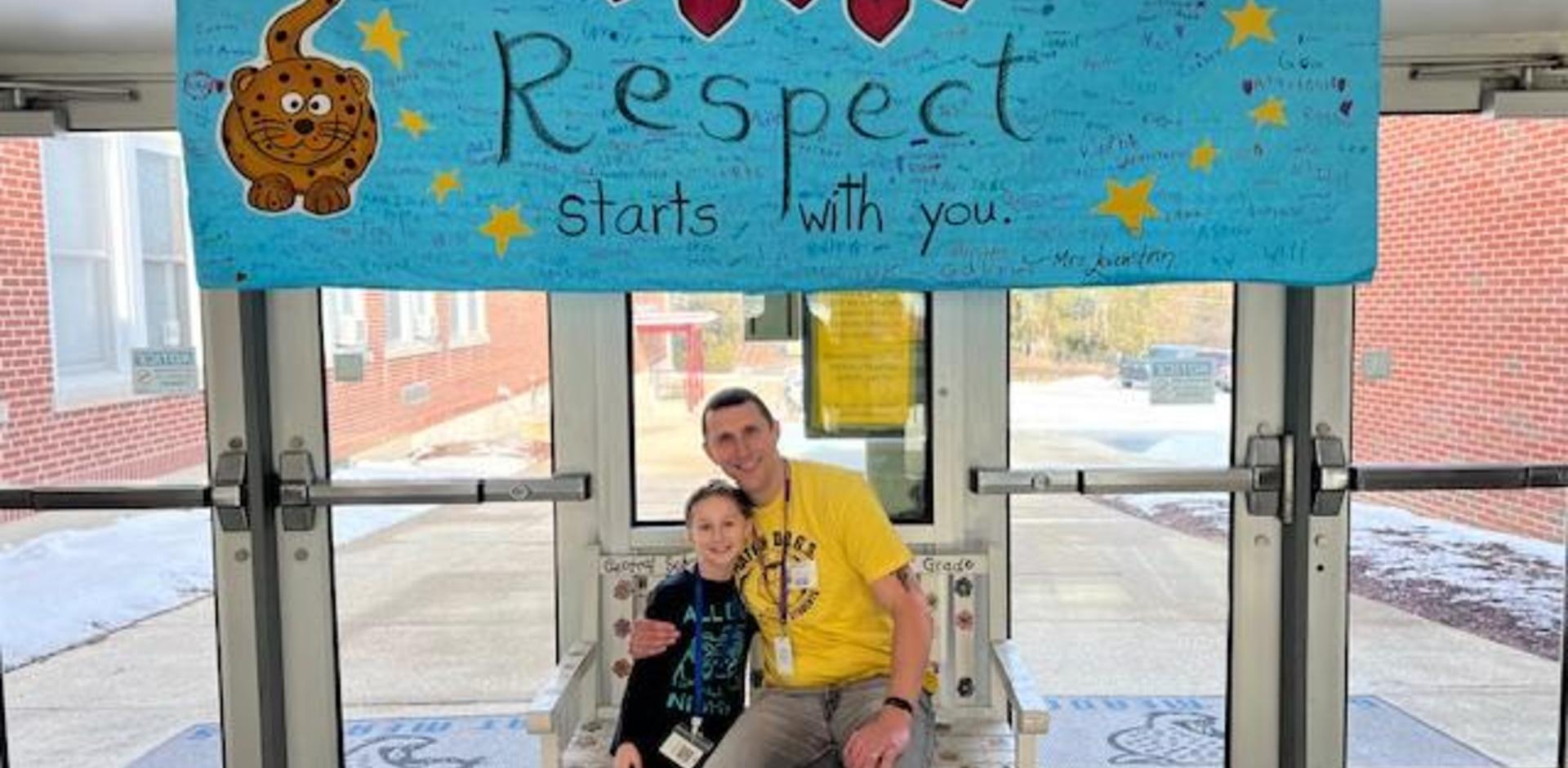 A man and a girl sitting under a 'Respect starts with you' banner at a school entrance.