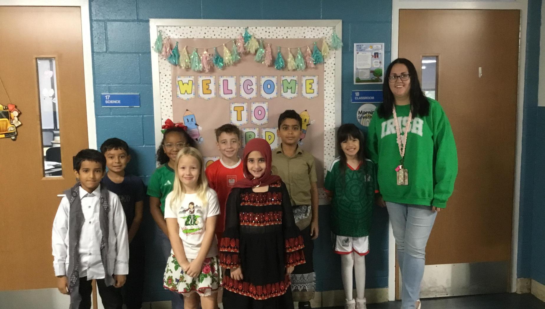 Diverse group of children in cultural dress smiles with a teacher near a 'Welcome' sign.