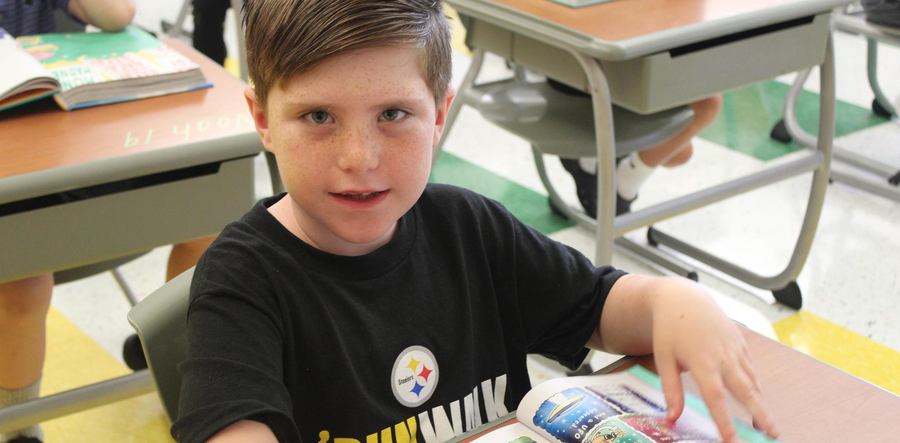 Smiling boy sitting at a desk, reading a colorful book.
