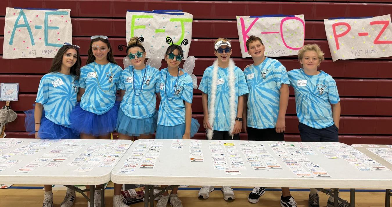 Seven children wearing blue shirts and accessories stand in front of decorated tables.