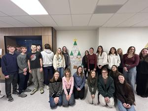 A group of students posing in front of a decorated Christmas tree on a wall.