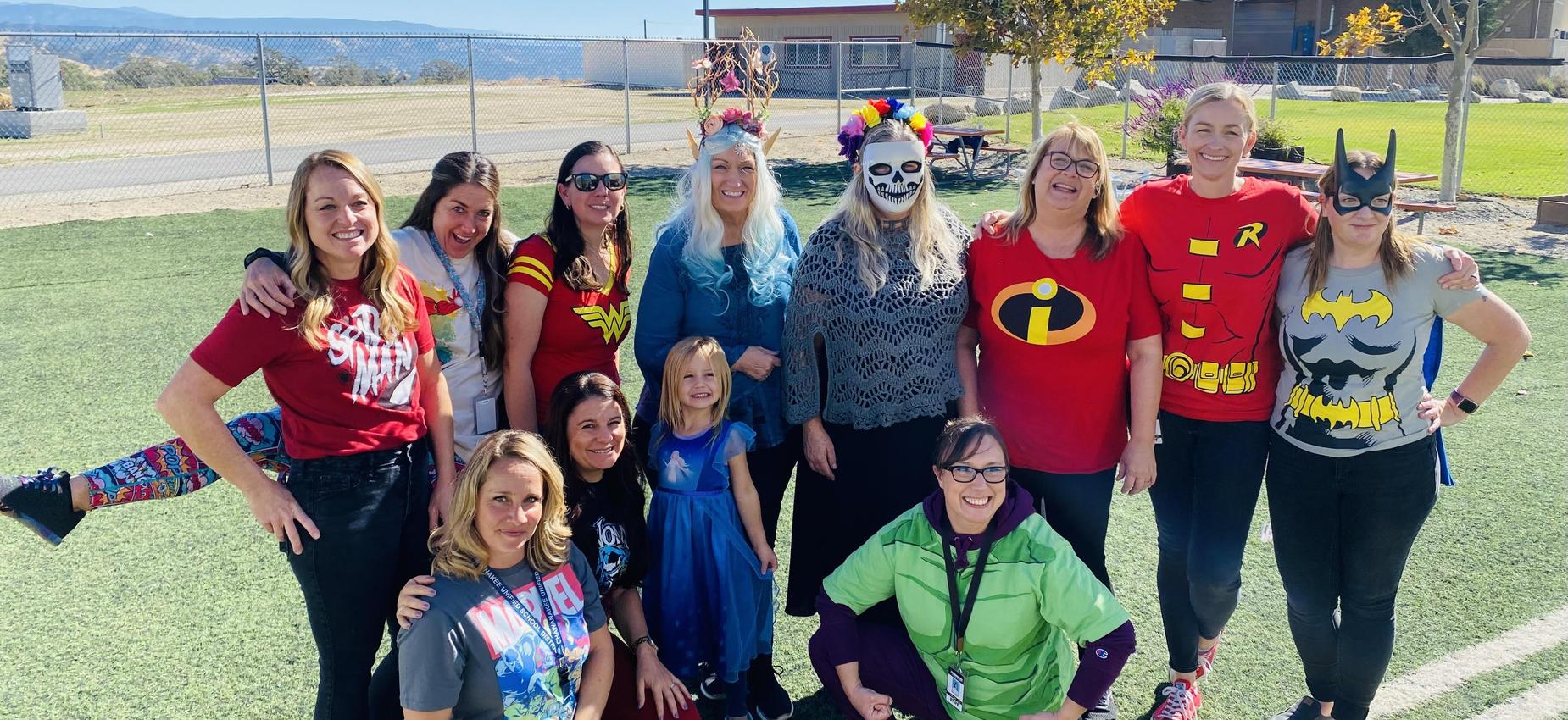 A group of women and girls in superhero costumes poses together outdoors.