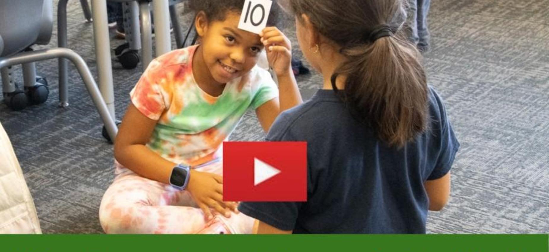 Two children interact in a classroom, one holding a number card on their forehead.