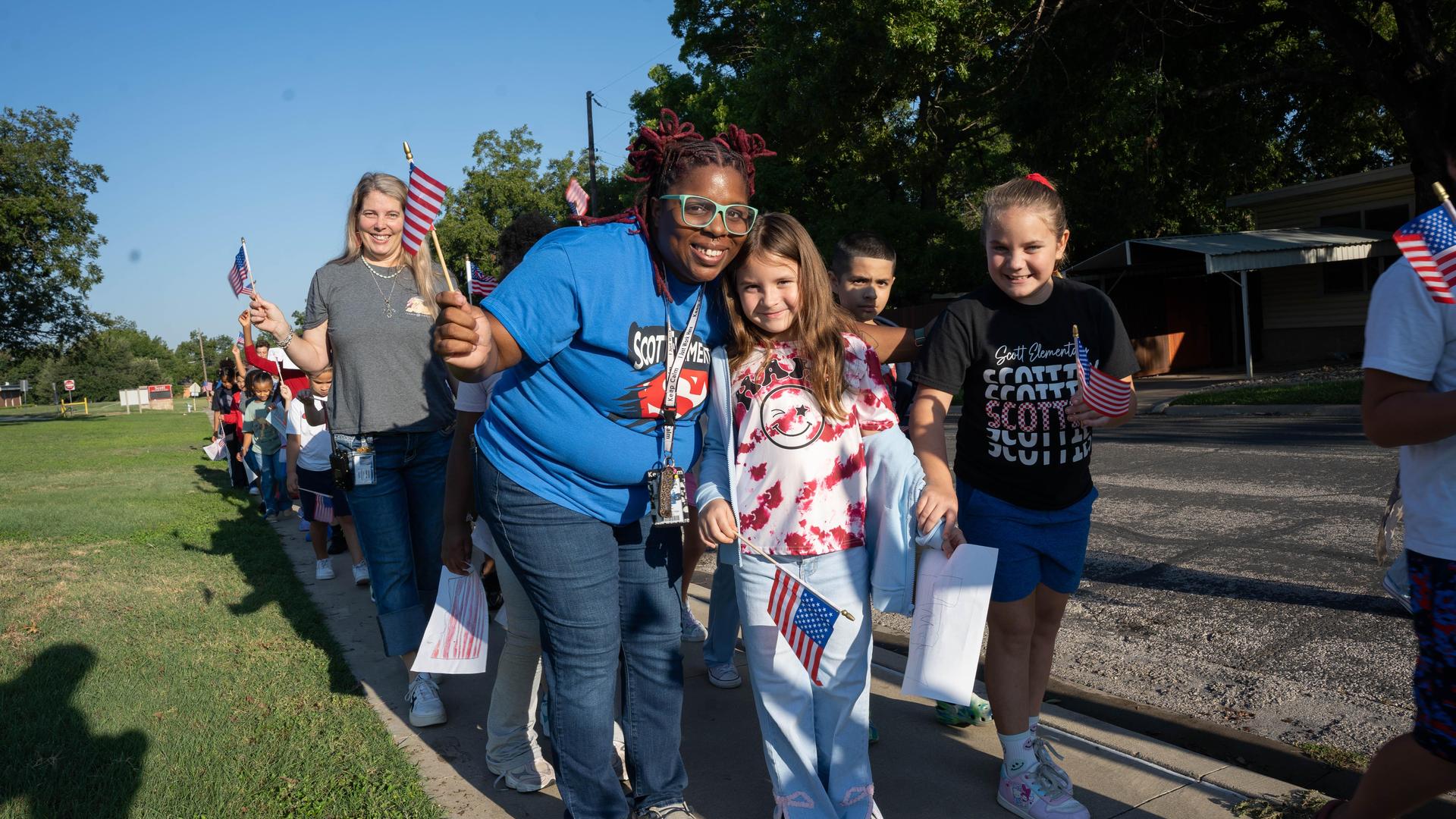 A group of children and adults walking with small American flags in a sunny park.