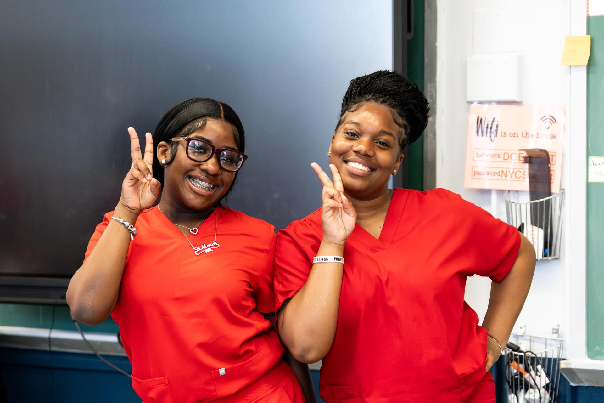 Two students in red scrubs pose playfully in a classroom, each making a peace sign with one hand.
