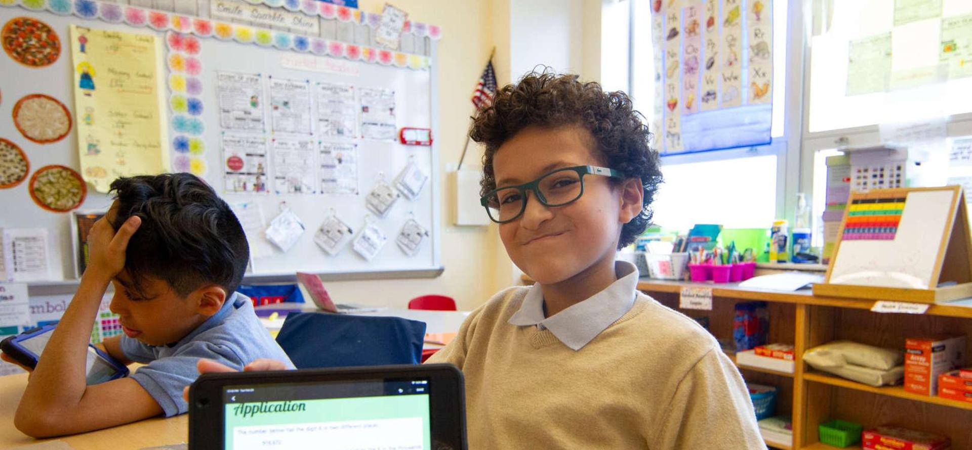 Boy smiling and showing a tablet in a classroom setting.