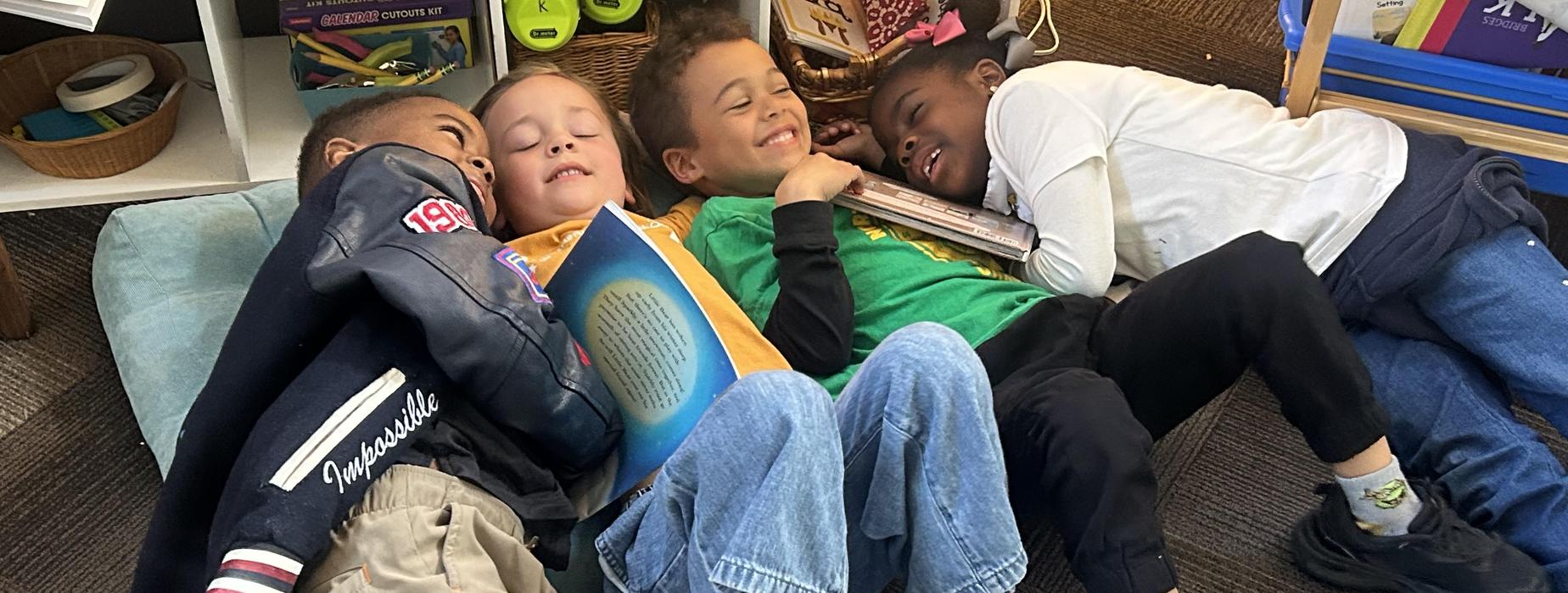 Four children lying close together on a carpet, reading and enjoying a story.