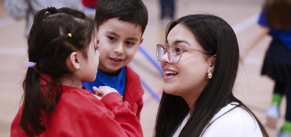 A woman smiles while interacting with two children in a gym setting.