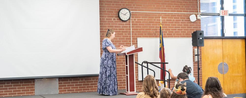 A woman in a dress speaks at a podium in front of an audience.