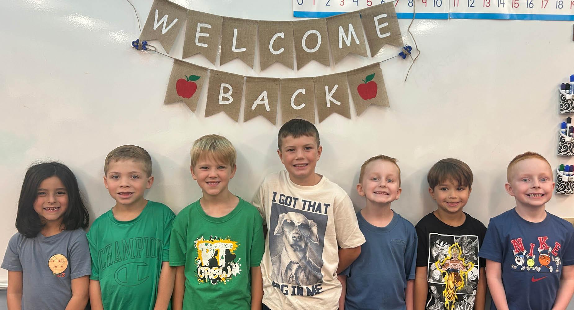 Group of children smiling under a 'Welcome Back' banner in a classroom.