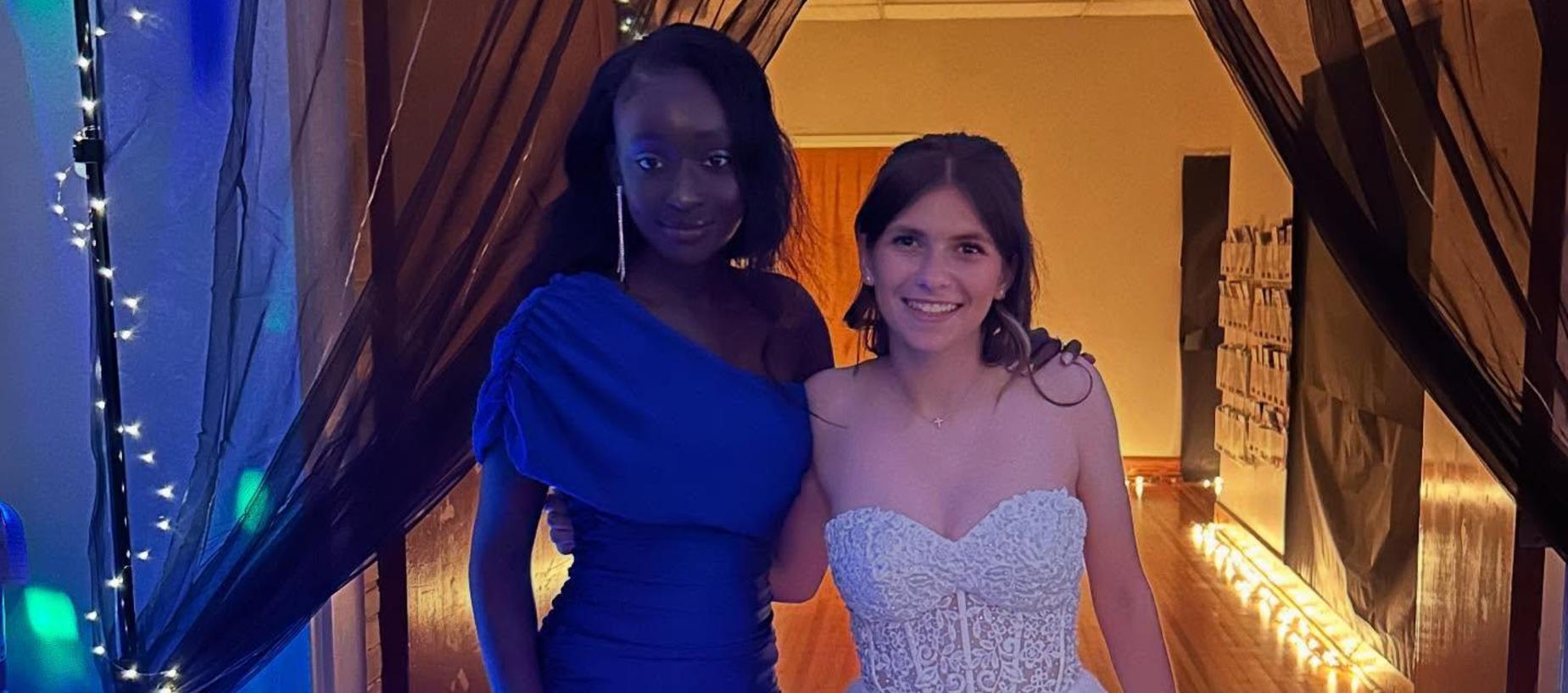 Two young women in formal dresses pose together in a softly lit hallway.