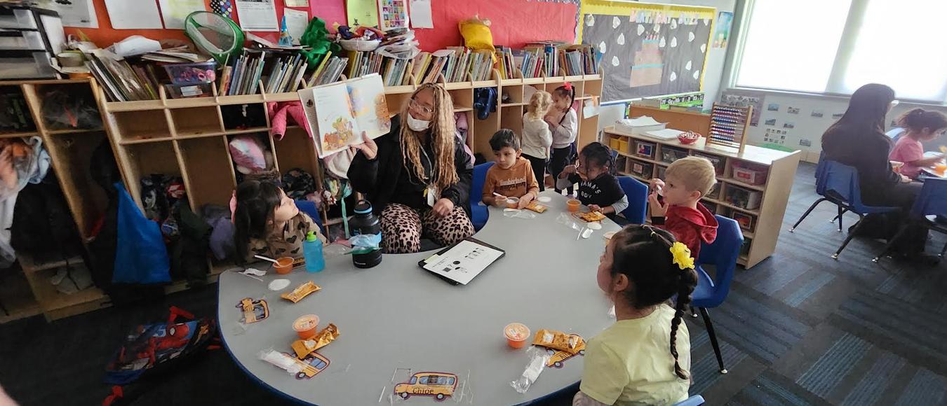 Children are sitting at a table eating fruit and crackers while a teacher is reading them a story.