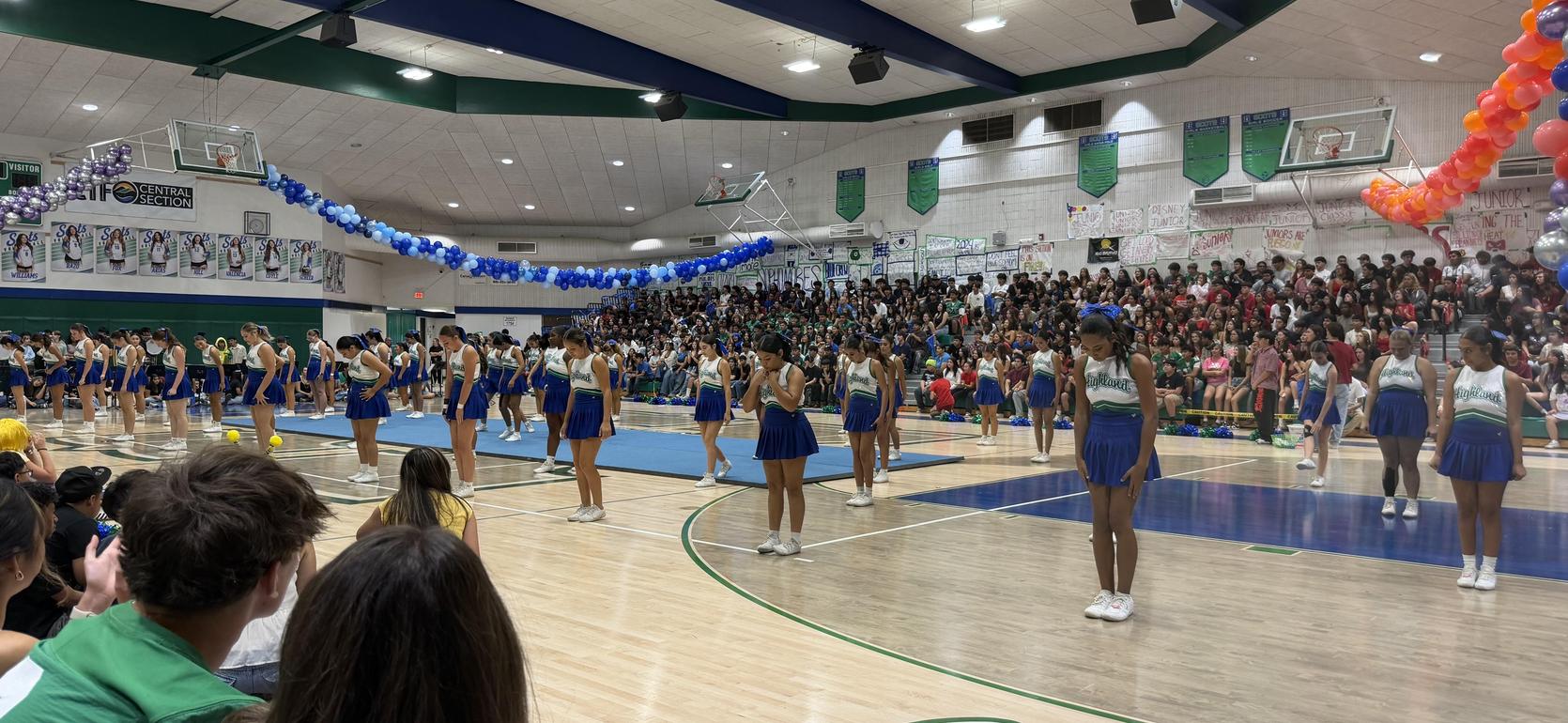 A school gymnasium filled with cheering students and performers in blue and green uniforms.