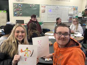 Two students holding drawings with friends in a busy classroom setting.