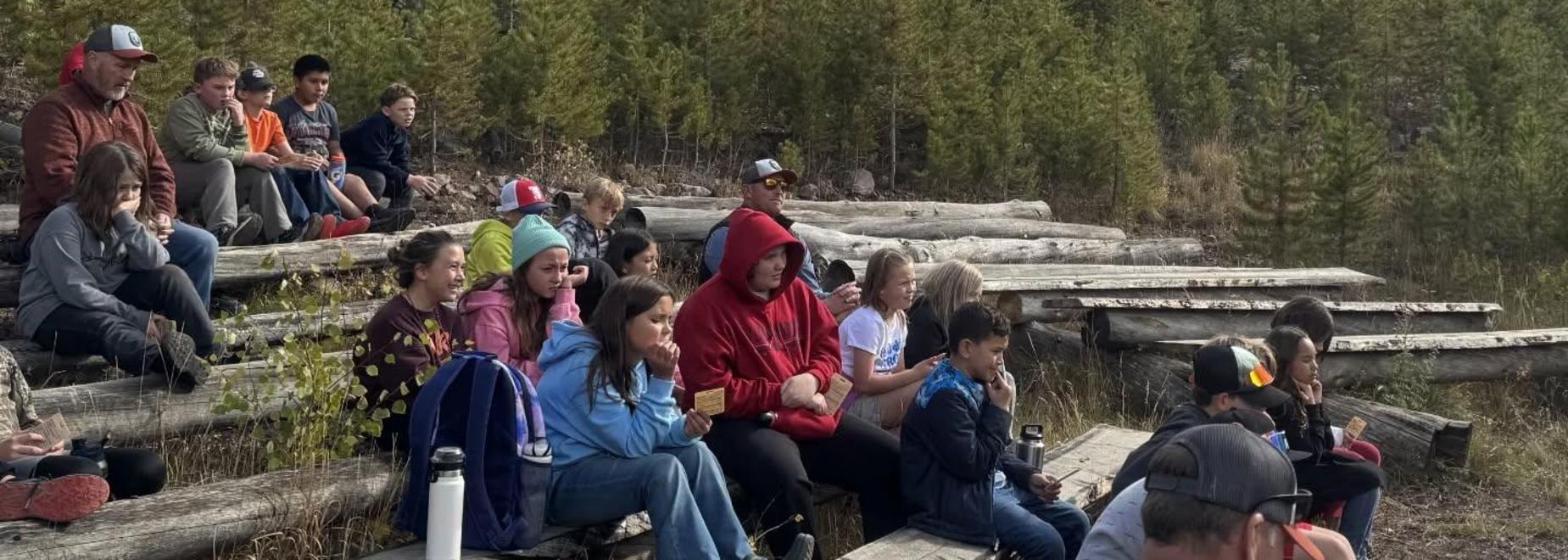 Group of children seated outdoors among trees during an activity.