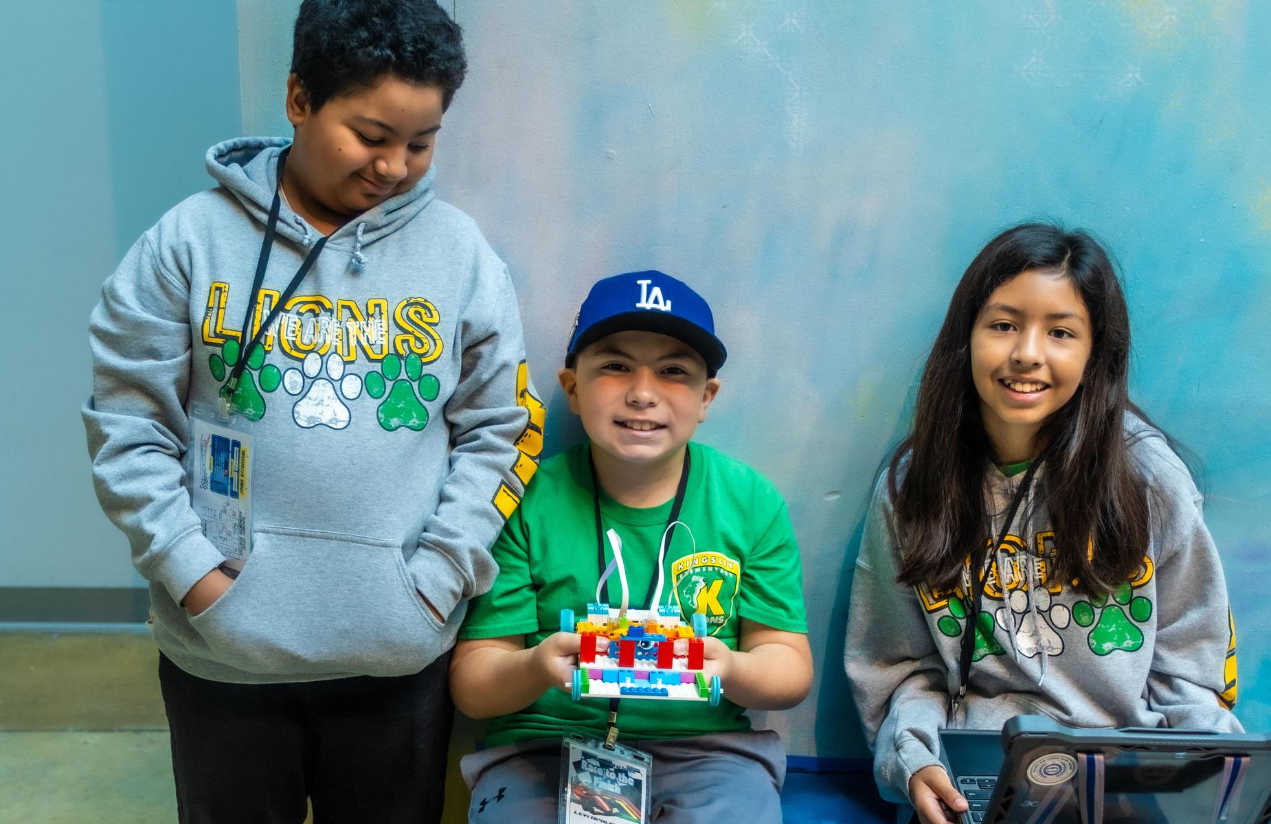 3 students, one in the middle is holding a lego robot and the other is loking down while another is holds a computer and smiles