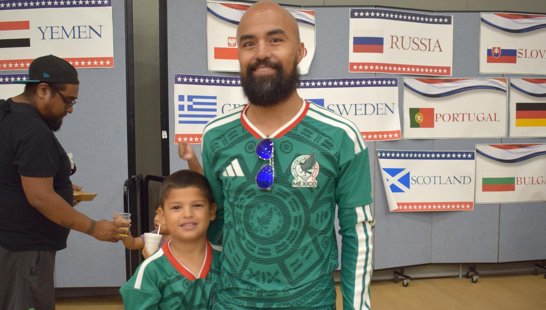 Man and boy in matching jerseys pose together at an event with flags in the background.