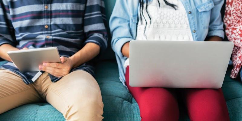 Two people sitting on a couch using a tablet and a laptop.