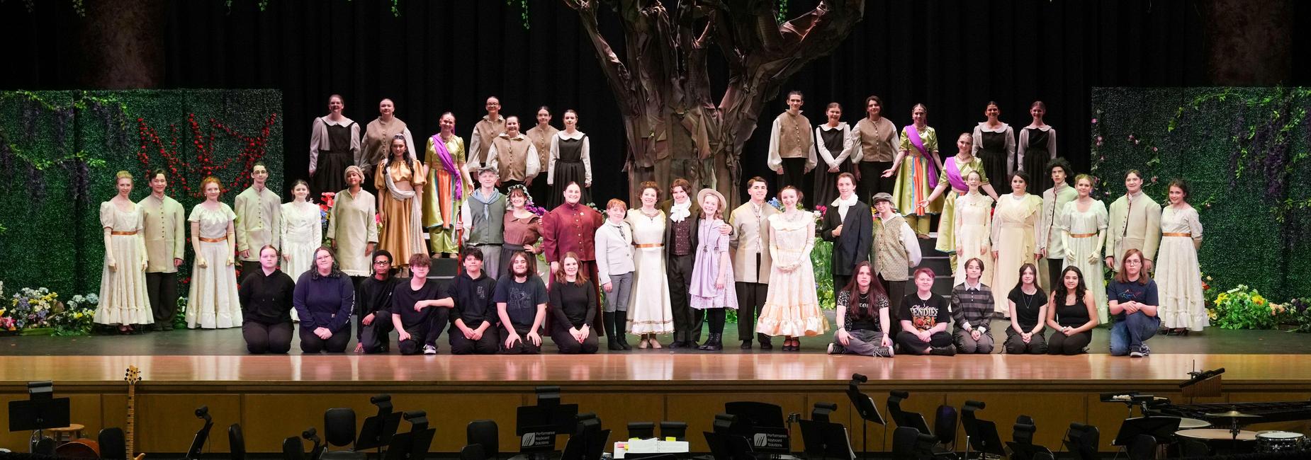 A large group of performers in costumes poses on stage with a tree backdrop cast of The Secret Garden.